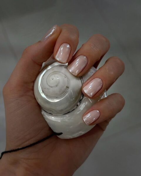 Perłowy manicure z efektem "sea shell nails" lub "oyster nails", inspirowany blaskiem macicy perłowej. Paznokcie o kwadratowym kształcie pokryte mleczną, różową bazą z opalizującym, chromowanym wykończeniem. Zadbane dłoń trzyma dużą muszlę ślimaka w kolorze bieli i perły, co podkreśla delikatny, oceaniczny trend w urodzie i jest inspiracją dla kobiet ceniących dyskretną elegancję i naturalne piękno.