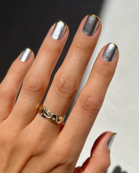Futurystyczny manicure z efektem "mixed metals" – paznokcie pomalowane są na metaliczne srebro i grafit z subtelnymi, złotymi akcentami na wolnym brzegu płytki (styl micro french lub złamany french). Ten modny trend w urodzie łączy chromowe wykończenie z elementami biżuteryjnymi. Ujęcie dłoni jest podkreślone przez gruby, złoty pierścionek z nieregularną fakturą, co stanowi inspirację dla kobiet ceniących awangardową elegancję i geometryczne detale.