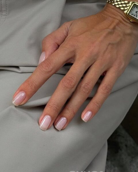 Perfekcyjnie zadbana dłoń z eleganckim, naturalnym manicure w stylu lipgloss nails lub mlecznej bazy, podkreślającym zdrowy wygląd i klasę. Na nadgarstku luksusowy, złoty zegarek na bransolecie. Dłoń spoczywa na spodniach z wysokiej jakości, beżowego materiału, co oddaje ducha minimalistycznego luksusu i świadomego doboru faktur w modzie i urodzie.