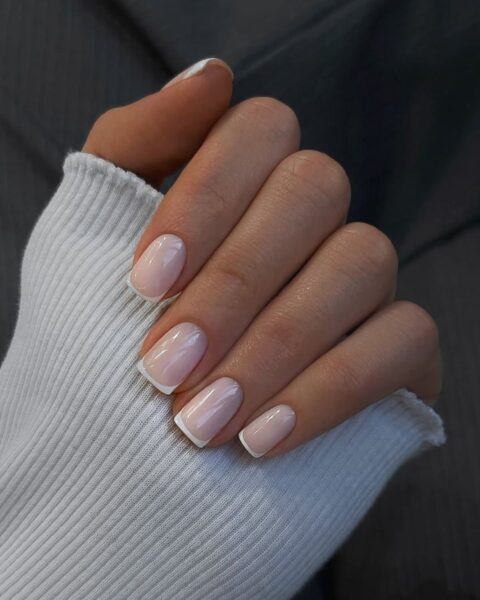 Ponadczasowy french manicure z perfekcyjnie wyrysowaną białą końcówką i mleczną, różową bazą. Ta klasyczna stylizacja paznokci o kształcie łagodnego kwadratu (squoval) jest kwintesencją dyskretnej elegancji i dbałości o detale w urodzie. Ujęcie dłoni w białym, kaszmirowym swetrze podkreśla trend quiet luxury i stanowi inspirację do stworzenia minimalistycznego, ale luksusowego looku.