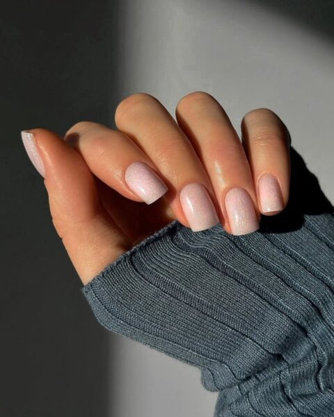 Delikatny manicure z efektem "glazed donut" lub efektem tafli w subtelnym, pudrowym różu, pięknie opalizujący w naturalnym świetle. Paznokcie o modnym, lekko kwadratowym kształcie i średniej długości, wpisujące się w estetykę clean girl aesthetic z nutą blasku. Dłoń w ciepłym, granatowym swetrze z widoczną fakturą materiału, co nadaje zdjęciu przytulny, jesienny klimat i podkreśla elegancję stylizacji.