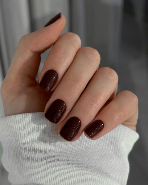 Elegancki, jesienny manicure w głębokim, lśniącym kolorze gorzkiej czekolady lub ciemnego bordo. Paznokcie o klasycznym, kwadratowym kształcie z zaokrąglonymi brzegami, symbolizujące ponadczasową elegancję. Ujęcie dłoni w białym, miękkim mankiecie swetra tworzy kontrast, który podkreśla intensywność koloru i stanowi idealną inspirację dla kobiet ceniących luksusowy minimalizm w urodzie.