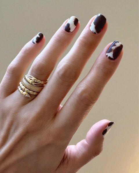 Modny manicure z wzorem krowich łat (cow print), czyli czarno-białym animal printem na paznokciach. To inspiracja dla kobiet, które szukają odważnego i awangardowego akcentu w urodzie. Uwagę zwraca także złoty pierścionek w kształcie węża na kilku palcach, podkreślający eklektyczny i modowy charakter całej stylizacji dłoni.