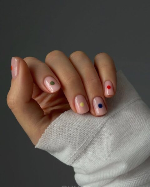 Kwadratowe paznokcie inspiracje: Manicure w stylu minimalistycznego pop-artu – pastelowa, różowa baza nude ozdobiona pojedynczymi, kolorowymi kropkami (dot nails) w kolorach: zielonym, żółtym, niebieskim i czerwonym. Subtelnie kwadratowe paznokcie są inspiracją dla kobiet z klasą, które lubią nowoczesne, dyskretne i geometryczne akcenty w urodzie. Dłoń ujęta w białym mankiecie swetra podkreśla współczesny minimalizm stylizacji.
