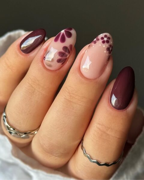 Elegancki, jesienny manicure na migdałowych paznokciach. Głęboki odcień burgundu przeplata się z beżowo-mleczną bazą, na której namalowano kwiatowe wzory w kolorze śliwkowym i bordowym oraz kropki. Stylizacja łączy bogate, jesienne kolory z delikatnym, kobiecym wzorem.