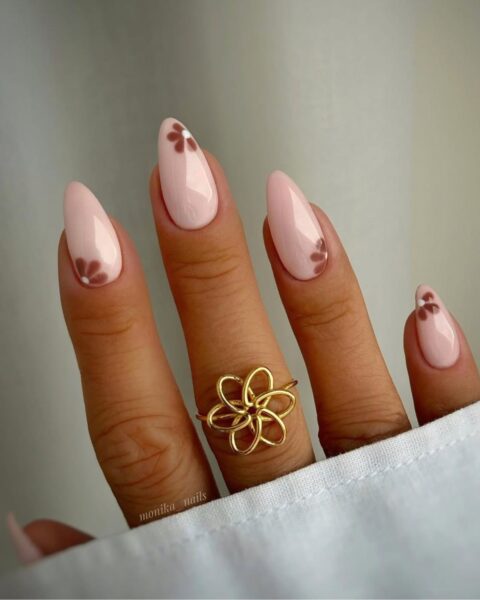Delikatny manicure w odcieniach beżu i brązu na migdałowych paznokciach. Jasnoróżowa (mleczno-różowa) baza ozdobiona jest małymi, brązowymi kwiatowymi wzorami z białym środkiem, umieszczonymi przy wolnym brzegu płytki. Dłoń zdobi duży, ażurowy złoty pierścionek w kształcie kwiatu. Stylizacja jest subtelna i elegancka.