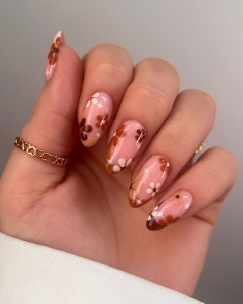 Żywy i modny manicure na jesień. Paznokcie w kształcie migdałków pokryte są mlecznoróżową bazą i ozdobione wzorem w brązowo-pomarańczowo-białe kwiaty, gdzieniegdzie z błyszczącymi, białymi akcentami przypominającymi gwiazdki. Wzór ma styl retro lub inspirowany latami 70. Modelka nosi na palcu cienki złoty pierścionek z motywem serc.