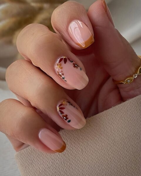 Delikatny i elegancki manicure na jesień. Paznokcie w kształcie zaokrąglonego migdałka utrzymane są w odcieniach nude i ozdobione jesiennymi akcentami. Niektóre paznokcie mają brązowo-złoty french, a paznokcie serdeczny i środkowy są udekorowane subtelnym motywem wianuszka z brązowych, bordowych i zielonych listków przy skórkach. Na palcu modelki widoczny jest cienki złoty pierścionek.
