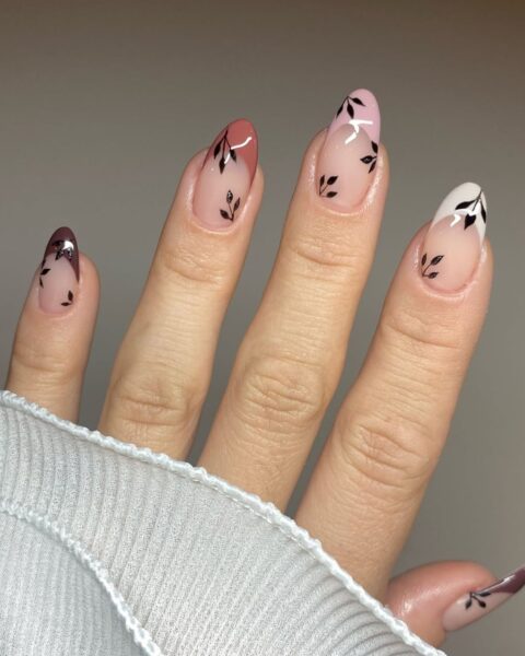 Elegancki manicure z jesiennym motywem liści na migdałowych paznokciach. Paznokcie mają bazę w kolorze nude lub mlecznego różu, a ich końcówki są pomalowane w odcieniach brązu, terakoty, bordo lub białego french. Na przezroczystej części płytki domalowano minimalistyczne, czarne gałązki z listkami. Stylizacja jest subtelna, ale wyrazista.