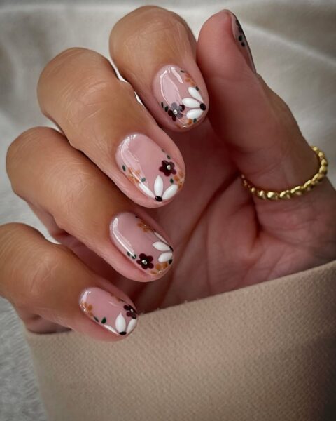 Uroczy, jesienny manicure na krótkich paznokciach w kształcie zaokrąglonego migdałka. Płytka pokryta jest mlecznoróżową (nude) bazą i ozdobiona wzorem w małe, stylizowane kwiaty w jesiennych kolorach: bordo, musztardowym, brązowym oraz białym z ciemnymi środkami. Modelka nosi na palcu cienki złoty pierścionek z kulkową fakturą. Stylizacja jest subtelna i kobieca.