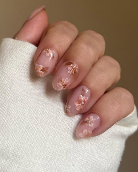 Delikatny, jesienny manicure na migdałowych paznokciach. Płytka jest pokryta półprzezroczystym, mlecznoróżowym lakierem i ozdobiona subtelnym wzorem w brzoskwiniowo-beżowe kwiaty i liście. Wzór przypomina akwarelę lub tusz, nadając stylizacji eteryczny i kobiecy charakter. Idealny manicure dla kobiet ceniących klasę i delikatność.