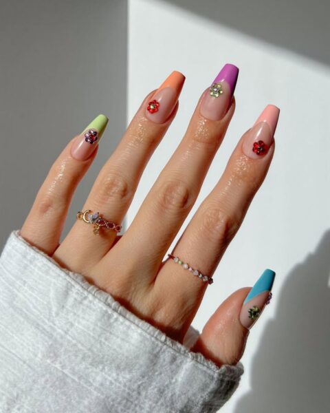 Dłoń w mankiecie białej koszuli prezentująca długi, kwadratowy manicure typu french z kolorowymi końcówkami. Każdy paznokieć ma końcówkę w innym, jaskrawym, neonowym kolorze (mięta, pomarańcz, fiolet, róż, turkus). Na każdym kolorowym fragmencie naklejony jest kryształkowy kwiatek w kontrastowym odcieniu. Na palcach widoczne są dwa delikatne, złote pierścionki z koralikami. Manicure jest ekstrawagancki i wakacyjny.