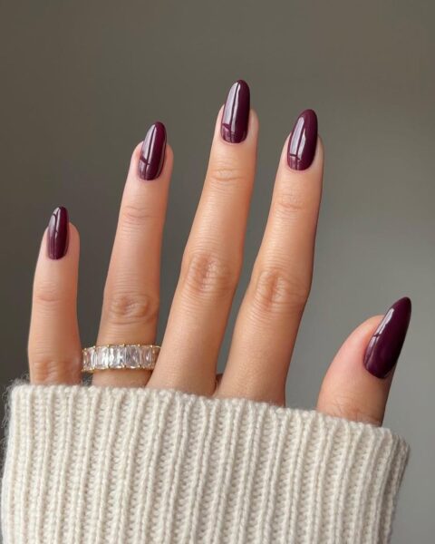 Zbliżenie na dłoń w mankiecie kremowego swetra, prezentującą długie paznokcie w kształcie migdała. Manicure jest wykonany błyszczącym, ciemnym lakierem w kolorze dojrzałej śliwki lub burgundu. Na palcu serdecznym widoczny jest szeroki, złoty pierścionek z rzędem prostokątnych, lśniących kamieni. Manicure jest elegancki i luksusowy.