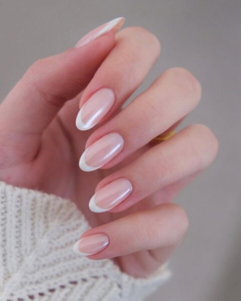 Manicure francuski na dłoni, w której paznokcie mają naturalny, migdałowy kształt. Płytka paznokcia jest pokryta delikatnym, perłowym lakierem w kolorze różowo-beżowym, a końcówki podkreślone cienką, białą linią. Dłoń częściowo zakrywa beżowy, dziergany sweter.