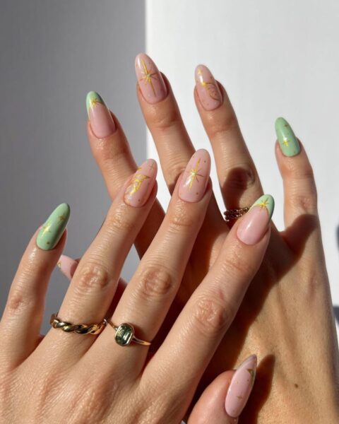Dwie dłonie prezentujące długi, migdałowy manicure inspirowany latami 90. oraz estetyką fairycore. Paznokcie są pomalowane na przemian jasnym, pudrowym różem i delikatną miętową zielenią. Na paznokciach widnieją złote zdobienia: gwiazdki, półksiężyce i motywy przypominające skrzydła motyla. Na palcach widoczne są złote pierścionki, w tym jeden z zielonym kamieniem i pleciony.