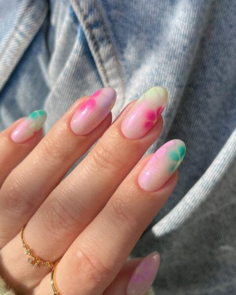 Zbliżenie na dłoń w mankiecie dżinsowej kurtki i miętowego swetra, prezentującą długie paznokcie w kształcie migdała. Manicure ma opalizującą, mleczną bazę, na której namalowano rozmyte, akwarelowe wzory kwiatów w intensywnych, pastelowych kolorach – różu i turkusowej zieleni. Na palcach widoczne są dwa delikatne, złote pierścionki, jeden z cyrkoniami, drugi z różowym kamieniem.