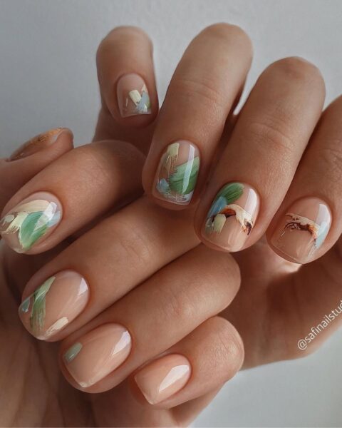 Manicure artystyczny w stylu abstrakcyjnego malarstwa. Krótka płytka w kształcie kwadratu/zaokrąglona, pokryta delikatną bazą nude/mleczną. Na każdym paznokciu znajduje się abstrakcyjny, malarski wzór w pastelowozielonych, beżowych i błękitnych barwach, przypominający akwarelowe pociągnięcia pędzla. To modna i kreatywna propozycja dla kobiet ceniących sztukę i oryginalne, subtelne zdobienia.