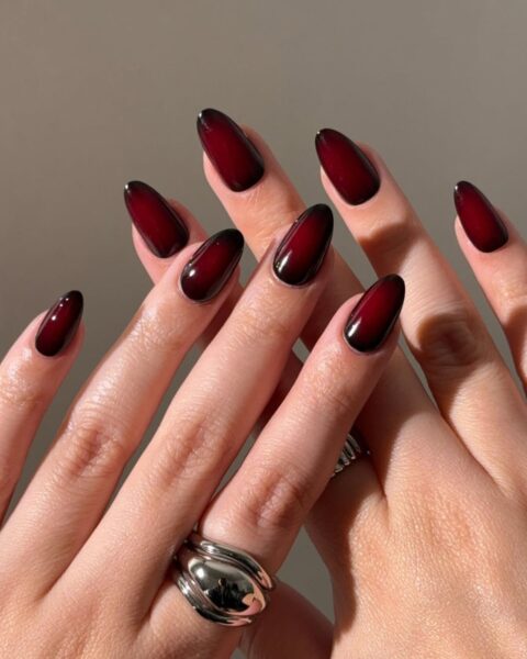 Gotycki manicure ombre (aura nails) na długich paznokciach o kształcie migdała/szpica. Stylizacja łączy głębokie, wampirze bordo (czerwień wina) z czarnym efektem dymu przy skórkach i na krawędziach. Błyszczące, mroczne i eleganckie paznokcie, idealne na październik, Halloween i jesienne wieczory.