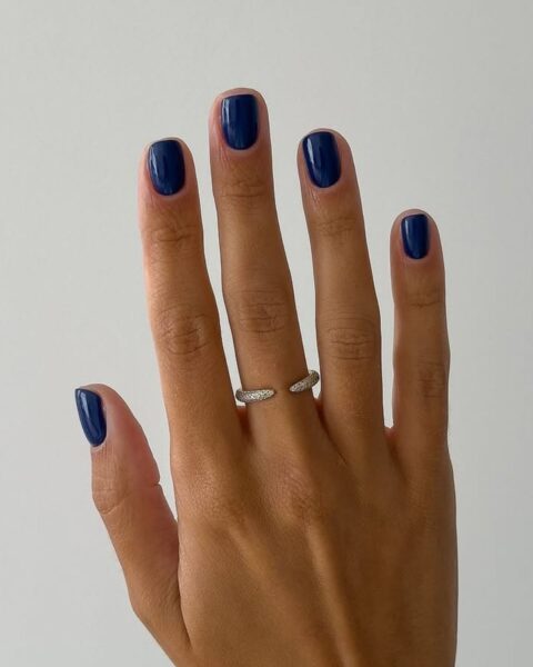 Elegancki manicure w kolorze granatu. Krótkie paznokcie o naturalnym, zaokrąglonym kształcie, pokryte błyszczącym, nasyconym lakierem w odcieniu atramentowym. Ten głęboki, wyrafinowany kolor stanowi luksusowy akcent, który pasuje zarówno do casualowych, jak i wieczorowych stylizacji, idealnie podkreślając dłonie z subtelną biżuterią.