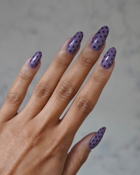 Jesienny manicure w groszki (polka dot nails). Długie paznokcie o modnym kształcie migdała, pomalowane błyszczącym, fioletowym (lawendowym/wrzosowym) lakierem. Zdobienie stanowią drobne, ciemnofioletowe kropeczki na całej płytce. Kreatywna i kobieca stylizacja paznokci, która nadaje jesiennym lookom uroku w stylu vintage/retro.