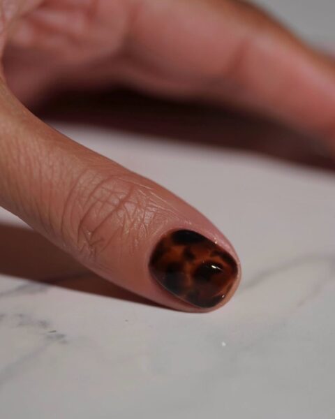 Jesienny manicure ze wzorem "szylkret" (tortoiseshell nails). Krótki paznokieć o naturalnym kształcie, pokryty błyszczącym wzorem imitującym skorupę żółwia – mieszanka głębokich brązów, czerni i ciepłych bursztynowych plam. To luksusowa i modna propozycja zdobienia paznokci, doskonale wpisująca się w jesienną estetykę i pasująca do złotych dodatków.