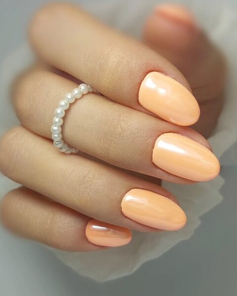 Delikatny manicure w odcieniu brzoskwini (peach nails) z subtelnym, perłowym połyskiem. Średniej długości paznokcie o kształcie migdała/owalnym w pastelowej, ciepłej tonacji. Słodki i elegancki kolor, idealny na lato lub jako akcent rozświetlający jesienne stylizacje. Na palcu widoczny pierścionek z perełek.