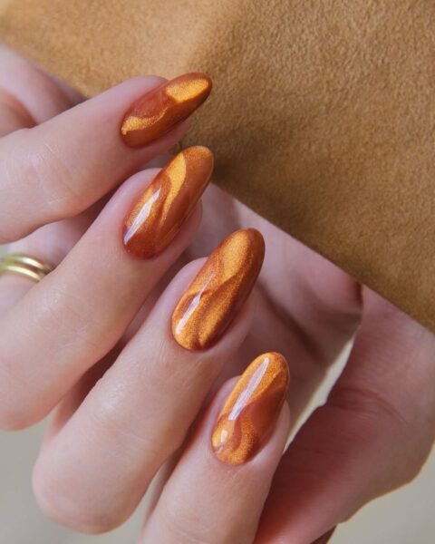 Jesienny, długi manicure w kształcie migdała (almond shape) z efektem kociego oka (magnetic nails). Paznokcie w głębokim, metalicznym odcieniu miedzi i rdzawej pomarańczy, tworzące dynamiczne, falujące wzory. Luksusowa i gorąca stylizacja paznokci, idealna na jesień, nawiązująca do kolorów dyni i liści.