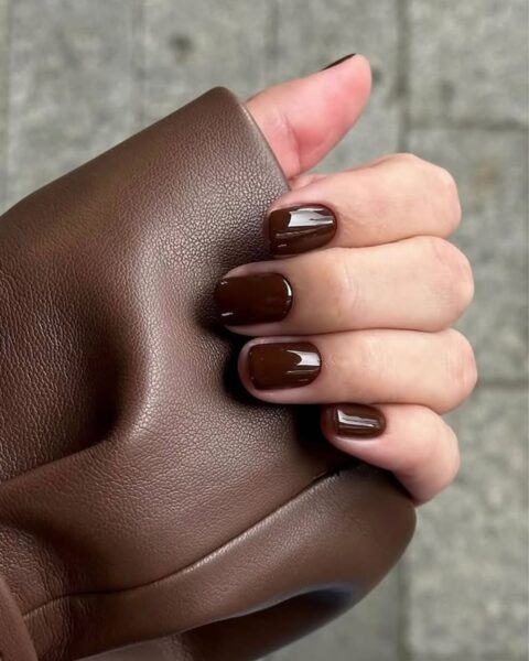 Manicure na pierwszą połowę października w stylu "Hot Chocolate Nails". Krótkie paznokcie o zaokrąglonym kształcie, pokryte błyszczącym, głębokim lakierem w kolorze gorzkiej czekolady/ciemnego brązu. Niezwykle elegancka i ponadczasowa stylizacja paznokci, idealna do skórzanych ubrań i jesiennych, ciepłych stylizacji.