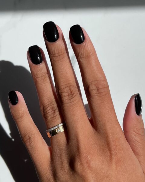 Elegancki, czarny manicure na krótkich paznokciach o zaokrąglonym, naturalnym kształcie. Paznokcie pomalowane są błyszczącym, głębokim lakierem w kolorze onyksu, który stanowi odważny, ale ponadczasowy akcent w stylizacji. Czarne paznokcie to modny wybór, który świetnie kontrastuje z opaloną skórą i złotą biżuterią.