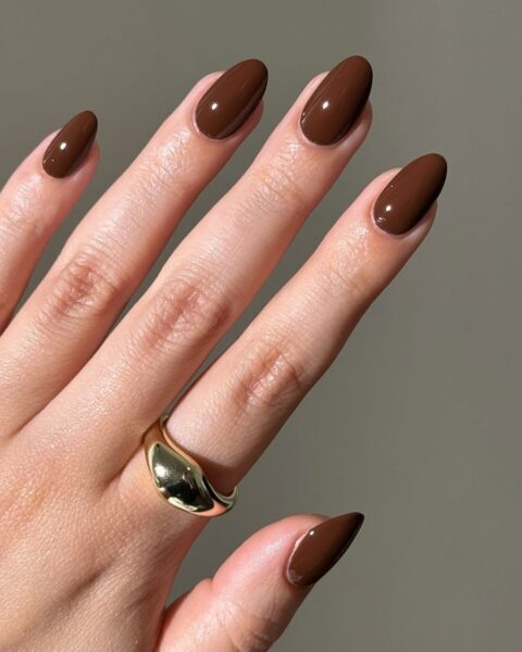 **Opis Alt:**Lśniący, jednolity manicure w jesiennych odcieniach: głębokim, ciepłym brązie (kolor gorzkiej czekolady) na długich, migdałowych paznokciach. Na palcu serdecznym widoczny jest duży, gładki, złoty pierścień w kształcie łezki.