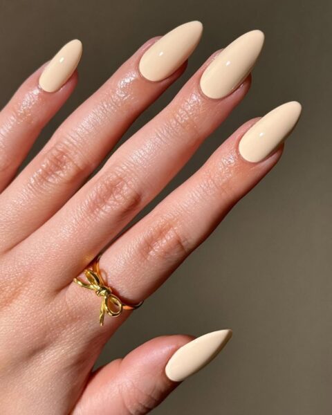 **Opis Alt:**Elegancki, kremowy manicure na długich, spiczastych (migdałowych/sztyletowych) paznokciach w lśniącym wykończeniu. Kolor to jasny beż lub waniliowy nudziak. Na palcu serdecznym widoczny delikatny złoty pierścionek w kształcie kokardki.
