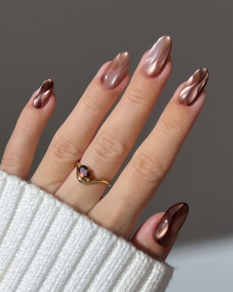 **Opis Alt:**Długie, migdałowe paznokcie z lśniącym manicure w jesiennych odcieniach brązu, miedzi i beżu. Na paznokciach widać efekt "płynnego metalu" i faliste wzory w stylu "swirl nails". Na palcu serdecznym złoty pierścionek z brązowym kamieniem.