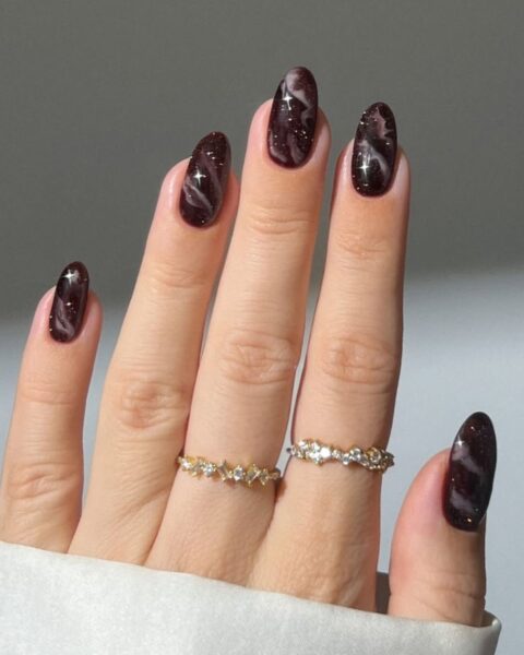 **Opis Alt:**Elegancki, owalny manicure w głębokim, jesiennym kolorze: ciemnym brązie z fioletową poświatą i drobnymi, brokatowymi drobinkami. Paznokcie zdobione są subtelnym, marmurkowym wzorem w odcieniu złamanej bieli. Na palcach serdecznym i środkowym dwa delikatne, złote pierścionki z cyrkoniami w kształcie gwiazdek.