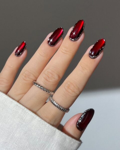 **Opis Alt:**Długie, owalne paznokcie z luksusowym manicure w kolorze głębokiej, wampirzej czerwieni (burgund lub wiśnia) z efektem kociego oka i srebrnym, falistym obramowaniem ("chrome border nails" lub "frame nails"). Na palcach środkowym i serdecznym proste, srebrne pierścionki o skręconej i żłobionej fakturze.