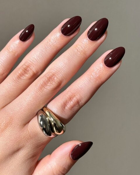 **Opis Alt:**Elegancki, błyszczący manicure na długich, migdałowych paznokciach, wykonany lakierem w kolorze głębokiego, jesiennego burgundu lub czekoladowej wiśni. Na palcu serdecznym widoczny duży, geometryczny pierścień w kolorach złota i srebra.