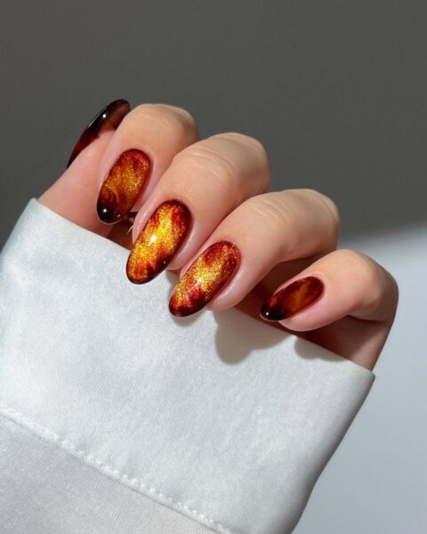 **Opis Alt:**Lśniący, owalny manicure z efektem kociego oka na długich paznokciach. Kolorystyka imituje jesienne płomienie lub bursztyn: połączenie głębokiego, ciemnego brązu/czerni na końcach z intensywnym, iskrzącym się pomarańczowo-złotym blaskiem w centrum. Manicure jest bardzo wyrazisty i luksusowy.