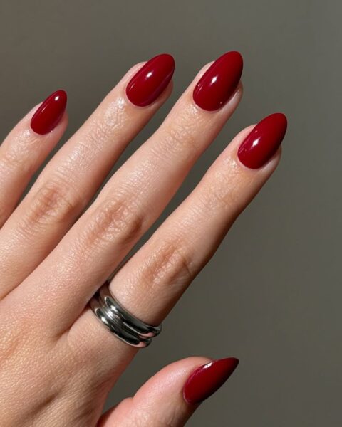 **Opis Alt:**Klasyczny, lśniący manicure na długich, migdałowych paznokciach, wykonany głęboką, elegancką czerwienią (burgund lub wiśniowa czerwień). Na palcu serdecznym widoczne są dwa proste, srebrne pierścionki. Manicure idealny na jesień i zimę.