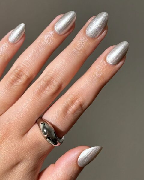 **Opis Alt:**Nowoczesny, metaliczny manicure na migdałowych paznokciach w kolorze perłowego srebra z efektem chromu. Lakiery dają efekt pionowej smugi światła. Na palcu serdecznym widoczny jest masywny, opływowy srebrny pierścień.