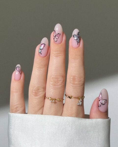 Manicure z motywem motyli – zbliżenie na zadbaną dłoń z paznokciami o kształcie owalnym lub migdałowym. Paznokcie pokryte są mlecznoróżową, transparentną bazą, na której umieszczono kolorowe, opalizujące naklejki lub stemple przedstawiające motyle. Motylki są w odcieniach różu, fioletu i czerni, dodając paznokciom eterycznego charakteru. Dłoń nosi delikatne, złote pierścionki.