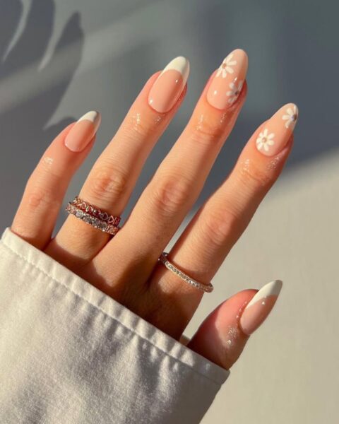 Manicure french z wiosennymi kwiatami – zadbana dłoń w białej koszuli na tle skąpanym w złotym świetle. Długie paznokcie o migdałowym kształcie pokryte są bazą w kolorze nude. Na kciuku i palcu wskazującym wykonano klasyczny, biały french, a na pozostałych paznokciach zamiast klasycznej końcówki namalowano białe, delikatne stokrotki. Dłoń zdobią dwa złote pierścionki.