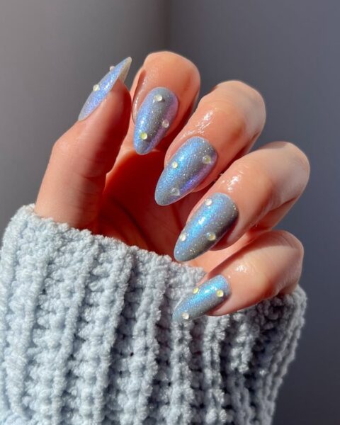 Manicure z efektem syrenki (holographic/chrome nails) – zbliżenie na dłoń ubraną w jasnoniebieski, wełniany sweter. Długie paznokcie o owalnym kształcie są pokryte niebiesko-fioletowym, opalizującym (holograficznym) lakierem, który mieni się brokatem. Każdy paznokieć ozdobiony jest kilkoma drobnymi, żółto-transparentnymi kryształkami (cyrkoniami), dodającymi paznokciom blasku.