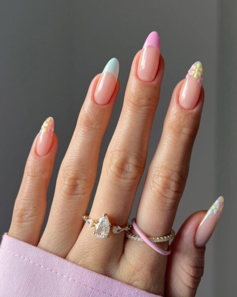 Manicure french w pastelowych kolorach – zbliżenie na zadbaną dłoń ubraną w różową bluzę, na tle neutralnej ściany. Długie, migdałowe paznokcie mają transparentną, różową bazę. Końcówki paznokci ozdobione są kolorowym frenchem, przy czym na palcu wskazującym jest on miętowo-niebieski, na środkowym różowy, a na pozostałych końcówka zastąpiona jest drobnymi, kolorowymi kwiatkami w odcieniach żółci, różu i zieleni. Dłoń zdobią dwa eleganckie pierścionki.
