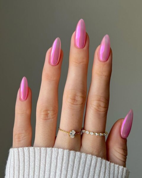 Chromowy manicure w kolorze fuksji – zbliżenie na dłoń w jasnym, wełnianym swetrze na neutralnym tle. Długie paznokcie w kształcie migdała są pokryte jaskrawym, chromowym lakierem w odcieniu intensywnego różu/fuksji, który odbija światło z metalicznym blaskiem. Dłoń zdobią dwa delikatne, złote pierścionki z kryształkami.