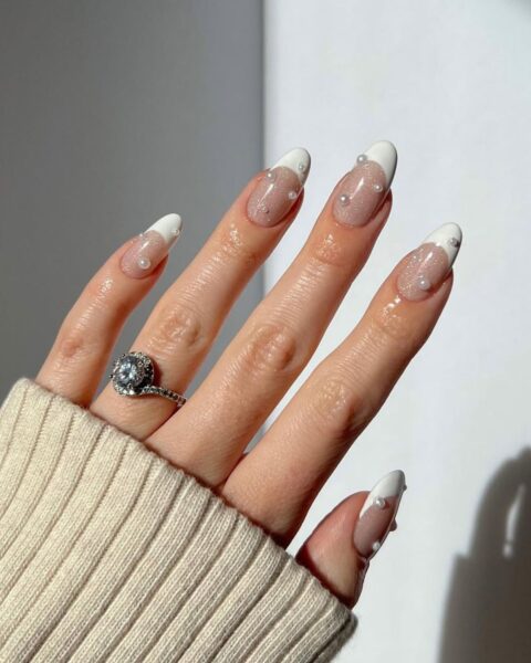 Manicure ślubny french z perłami – zbliżenie na dłoń w jasnym, beżowym swetrze, na której widać pierścionek zaręczynowy. Długie paznokcie w kształcie migdała mają klasyczny french manicure z wyraźną, białą końcówką. Baza paznokci jest transparentna i lekko brokatowa. Na środku każdego paznokcia (u nasady lub wzdłuż końcówki) umieszczone są małe, białe perełki 3D.