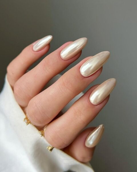 Manicure Vanilla Chrome – zbliżenie na zadbaną dłoń z długimi paznokciami o kształcie migdała. Paznokcie pokryte są opalizującym, chromowym lakierem w kolorze jasnego beżu lub wanilii, dającym perłowy, metaliczny blask. Dłoń jest ubrana w mankiet białej koszuli, a jej naturalny, elegancki wygląd wpisuje się w estetykę "clean girl aesthetic".