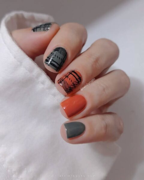 Nowoczesny i asymetryczny manicure w kratkę na krótkich, zaokrąglonych paznokciach. Paznokcie pomalowane są na jesienne kolory: matową szarość (mały palec i część kciuka) i ceglany pomarańcz (palec serdeczny). Paznokieć środkowy ma czarno-pomarańczową kratę, a wskazujący – szaro-czarną kratę. Wzory kraty są abstrakcyjne i graficzne. Dłoń częściowo ukryta w mankiecie białej koszuli lub swetra. Stylowa inspiracja na jesienny nail art.