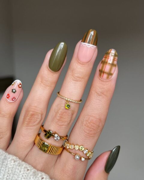 Luksusowy, jesienny manicure w kratkę na długich paznokciach o kształcie migdała, łączący kilka trendów. Paznokieć środkowy pokryty jest klasyczną kratą (tartan) w odcieniach beżu, różu i brązu. Paznokcie mały i kciuk pomalowane są na elegancką, głęboką zieleń oliwkową, a wskazujący ma wzór french manicure z zielonymi końcówkami, odcięty złotym paskiem i falbanką. Paznokieć serdeczny ma delikatny wzór kwiatowy na przezroczystej bazie. Dłoń ozdobiona złotymi pierścionkami z zielonymi i żółtymi kamieniami szlachetnymi, a w tle widać jasny, wełniany sweter. Stylowa, wielowymiarowa inspiracja na jesienny nail art.