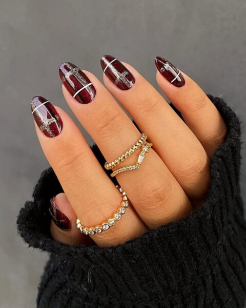 Glamour manicure w kratkę na długich, migdałowych paznokciach, idealny na jesień lub zimę. Wzór kraty jest wykonany z cienkich, białych linii oraz błyszczących, brokatowych linii w kolorze szampańskiego złota/srebra, na tle głębokiej, bordowej lub ciemnowiśniowej bazy. Dłoń ozdobiona jest złotą biżuterią – dwoma pierścionkami (jeden w kształcie chevronu, drugi z rzędem kryształków) i częściowo otulona mankietem czarnego, dzianinowego swetra.