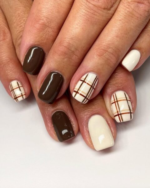 Manicure hybrydowy idealny na jesień, z krótkimi, kwadratowymi paznokciami, utrzymany w ciepłej, czekoladowej i kremowej kolorystyce. Niektóre paznokcie są pomalowane na jednolity, lśniący, głęboki brąz lub kremową biel. Pozostałe paznokcie mają wzór manicure w kratkę (plaid) w odcieniach brązu i beżu na kremowej bazie. Elegancka i klasyczna, czekoladowa inspiracja na paznokcie.