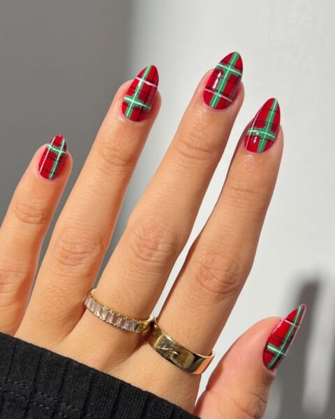 Mocny akcent świątecznego manicure w kratkę (tartan) utrzymany w klasycznych barwach Bożego Narodzenia: lśniącej czerwieni i zieleni. Wszystkie paznokcie o wydłużonym, szpiczastym kształcie migdała, pokryte są wzorem szkockiej kraty z cienkimi, zielonymi liniami na czerwonym tle. Dłoń ozdobiona złotymi pierścionkami, w tym jednym z rzędem kryształków, częściowo widoczna w mankiecie czarnego swetra. Idealna inspiracja na świąteczny nail art.